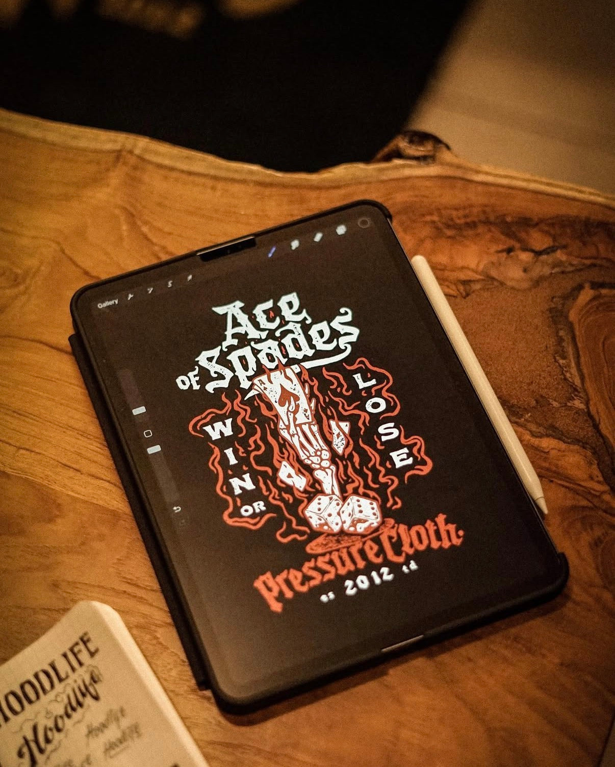 Tablet mit digitaler Zeichnung eines brennenden Ass Pik mit Würfeln und Schriftzug Pressure Cloth auf Holzuntergrund